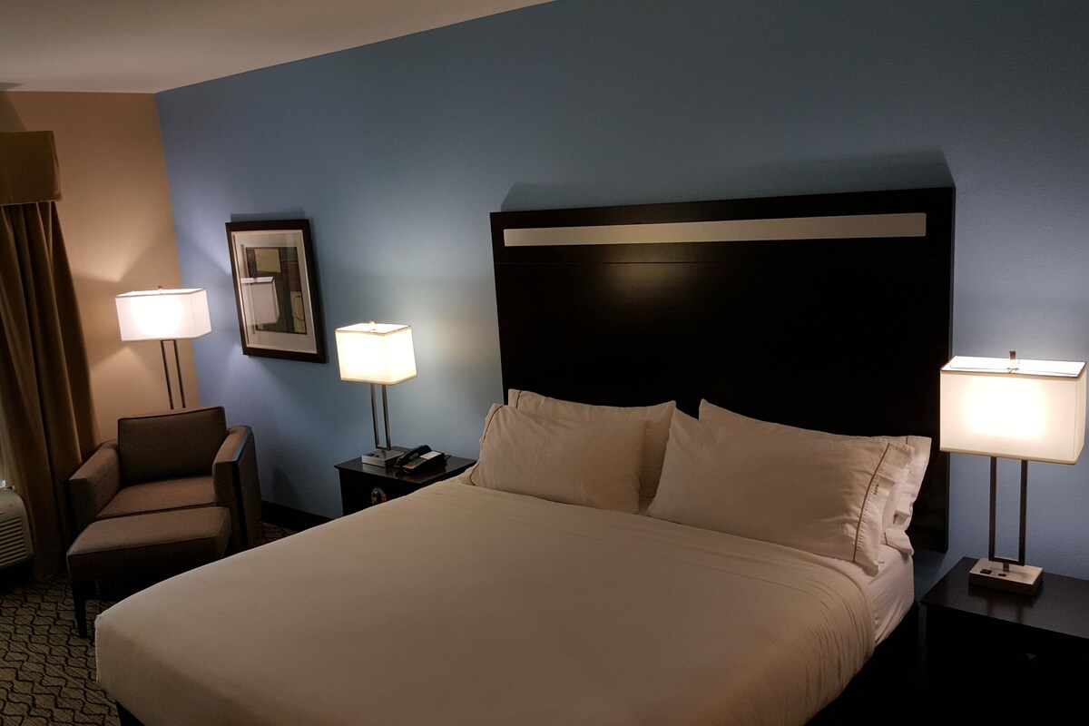 Habitación de hotel con cama, dos lámparas de noche, una silla, mesita de noche, cuadro enmarcado y ventana con cortinas.