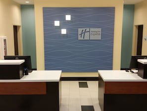 Zona de recepción con pared azul que presenta un diseño ondulado y el logotipo de Holiday Inn Express. Hay dos mostradores de recepción con computadoras y un piso de baldosas.