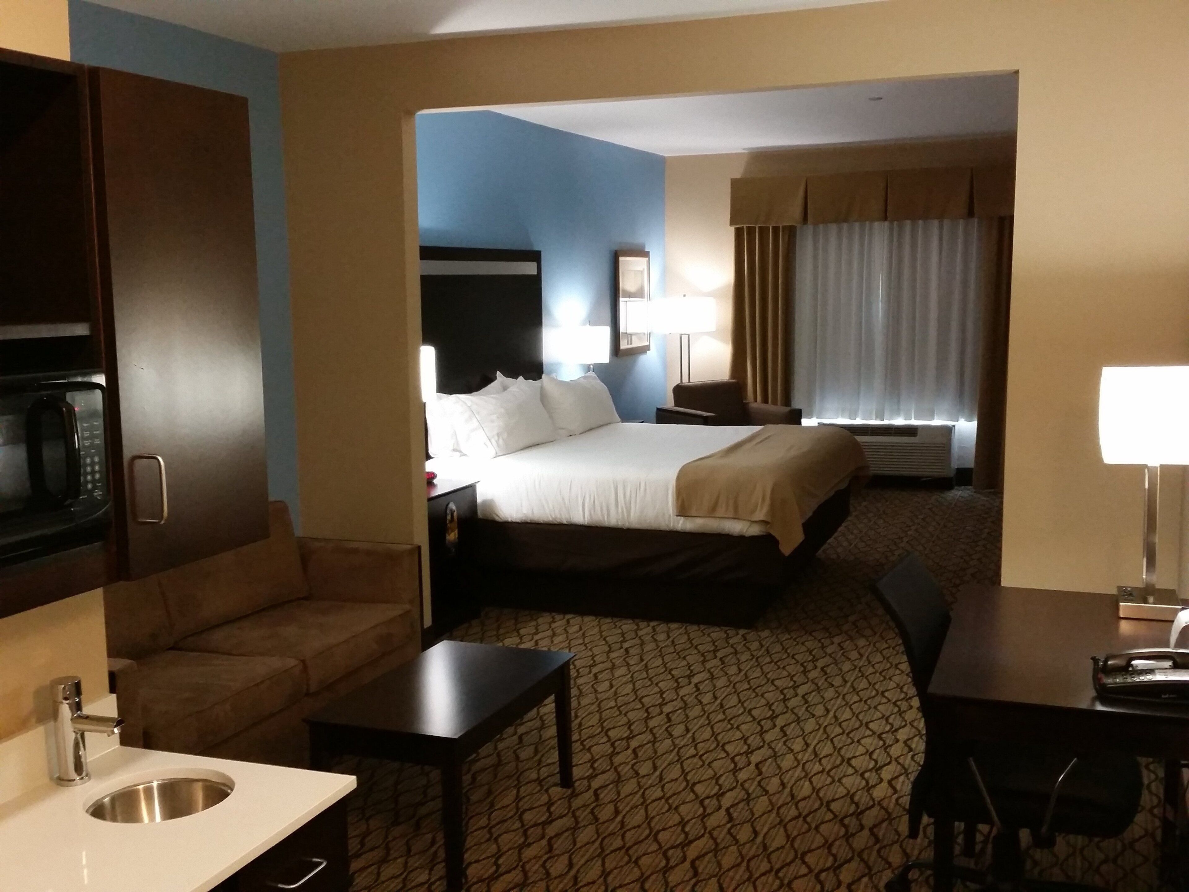 A hotel room with a bed, a desk, a sofa, a microwave, a sink, and a window with curtains.