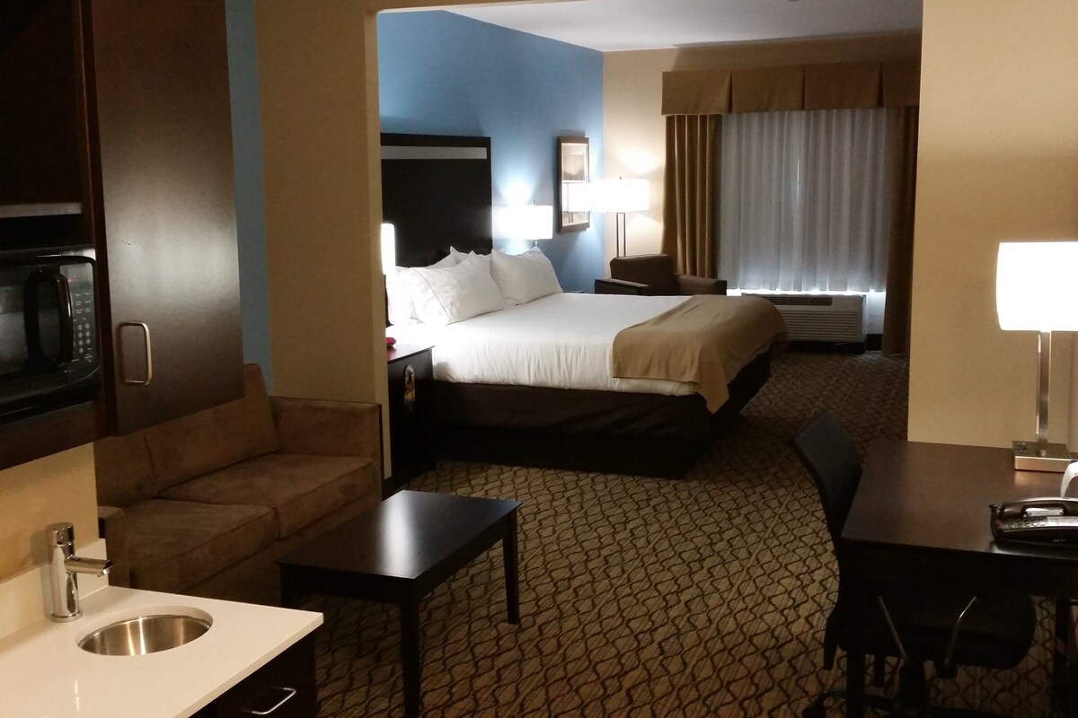 Habitación de hotel con cama, escritorio, sofá, microondas, lavamanos y ventana con cortinas.