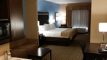 A hotel room with a bed, a desk, a sofa, a microwave, a sink, and a window with curtains.