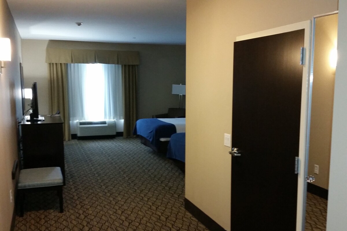 Una habitación de hotel con cama, escritorio con silla, ventana con cortinas y un equipo de aire acondicionado.