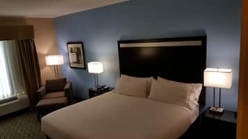 Une chambre d’hôtel avec un grand lit, deux lampes de chevet, une chaise, une table de chevet, un tableau encadré et une fenêtre avec des rideaux.