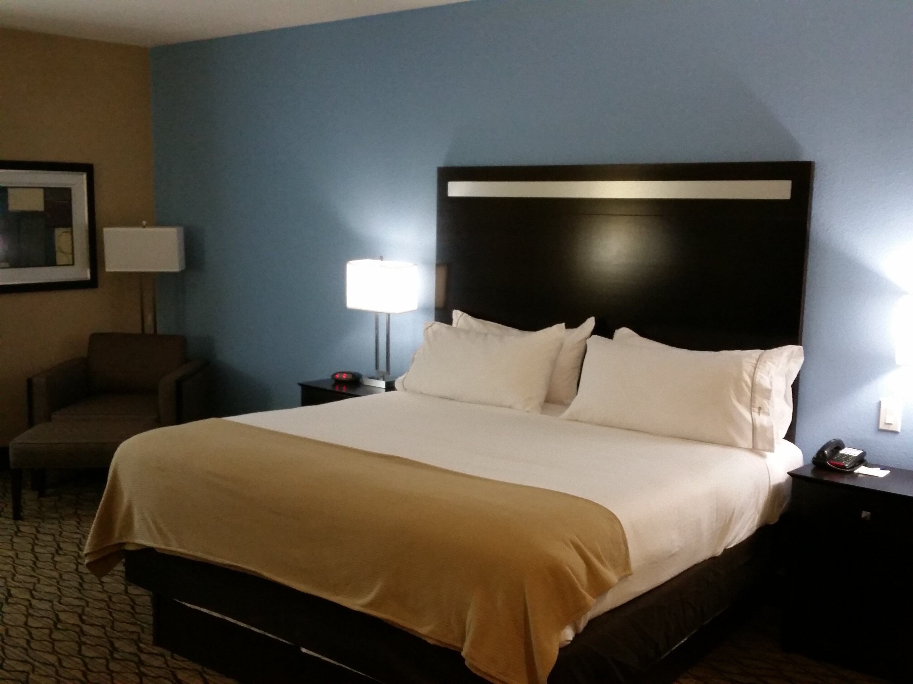 Una habitación de hotel con una cama grande, una mesita de noche con una lámpara, una silla y un cuadro en la pared.