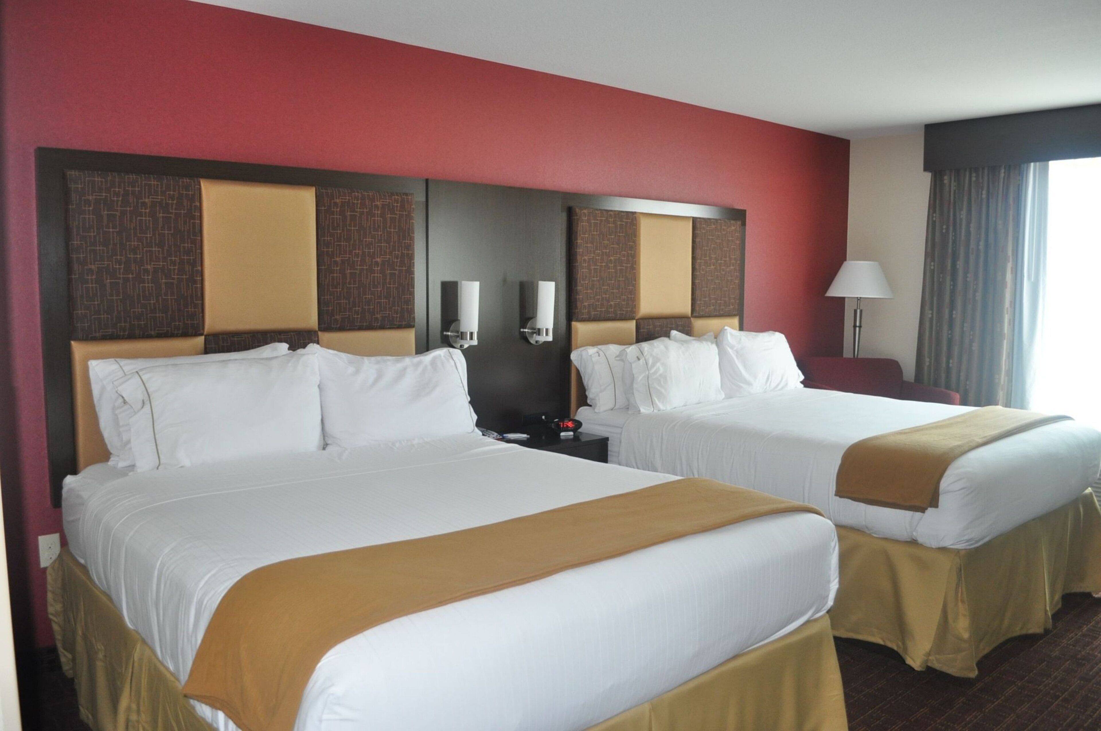 Habitación de hotel con dos camas, una mesita de noche, una lámpara y un ventanal con cortinas.