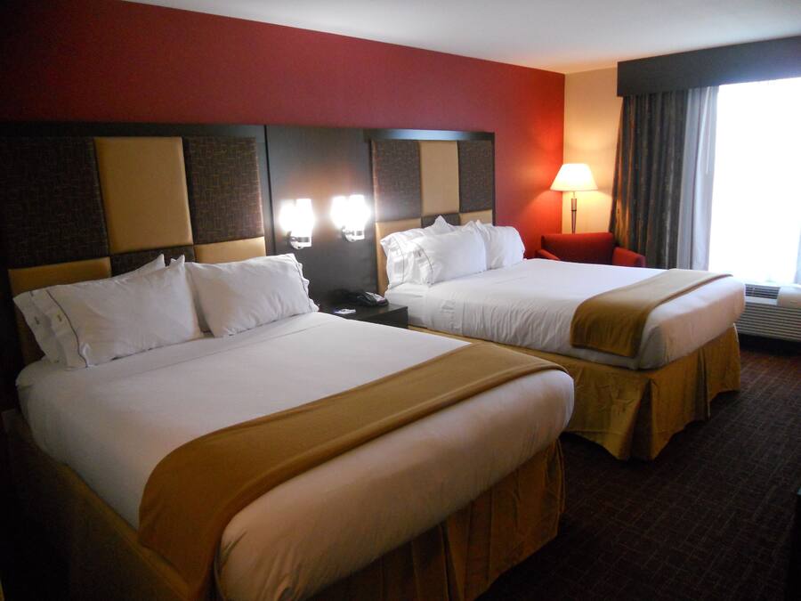 A hotel room with two beds, a red accent wall, a nightstand with a lamp, and a window with curtains.
