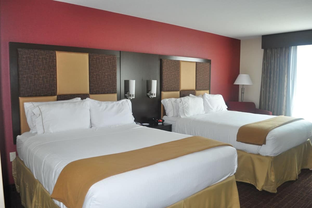 Une chambre d’hôtel avec deux lits, chacun agrémenté de draps blancs et d’un plaid marron. Il y a deux tables de chevet avec des lampes et une horloge. La chambre est dotée d’un mur d’accent rouge et d’une grande fenêtre avec des rideaux.