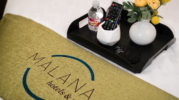 Un lit d’hôtel avec un plateau contenant une bouteille, une télécommande et un vase avec des fleurs. L’oreiller du lit porte le nom et le logo de l’hôtel.
