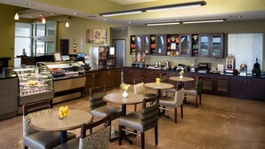 A restaurant with round tables, cushioned chairs, and a display case with food items. - Malana Hotels & Suites (Cotulla)