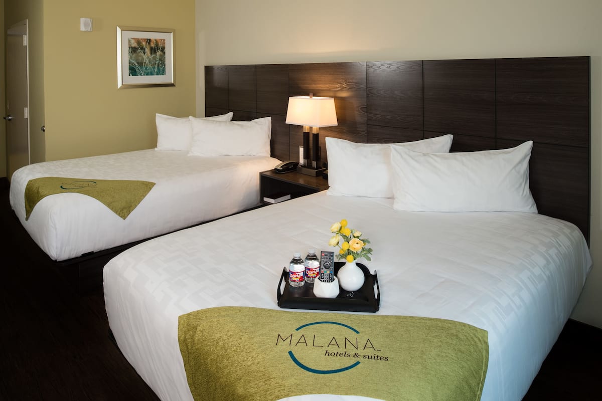 A hotel room with two beds, a nightstand with a lamp, a tray with toiletries, and a Malana hotels & suites logo on the bedspread.
