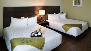 A hotel room with two beds, a nightstand with a lamp, a framed picture on the wall, and a tray with toiletries on the bed. - Malana Hotels & Suites (Cotulla)