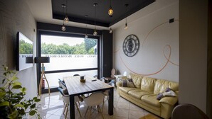 A modern living room with a wooden dining table, a beige sofa, and a large window offering a view of greenery. - Hotel Piccolo Allamano (Grugliasco)
