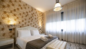 A bedroom with a brick-patterned wall, a large bed with white bedding, a bench at the foot of the bed, and a window with sheer curtains. - Hotel Piccolo Allamano (Grugliasco)