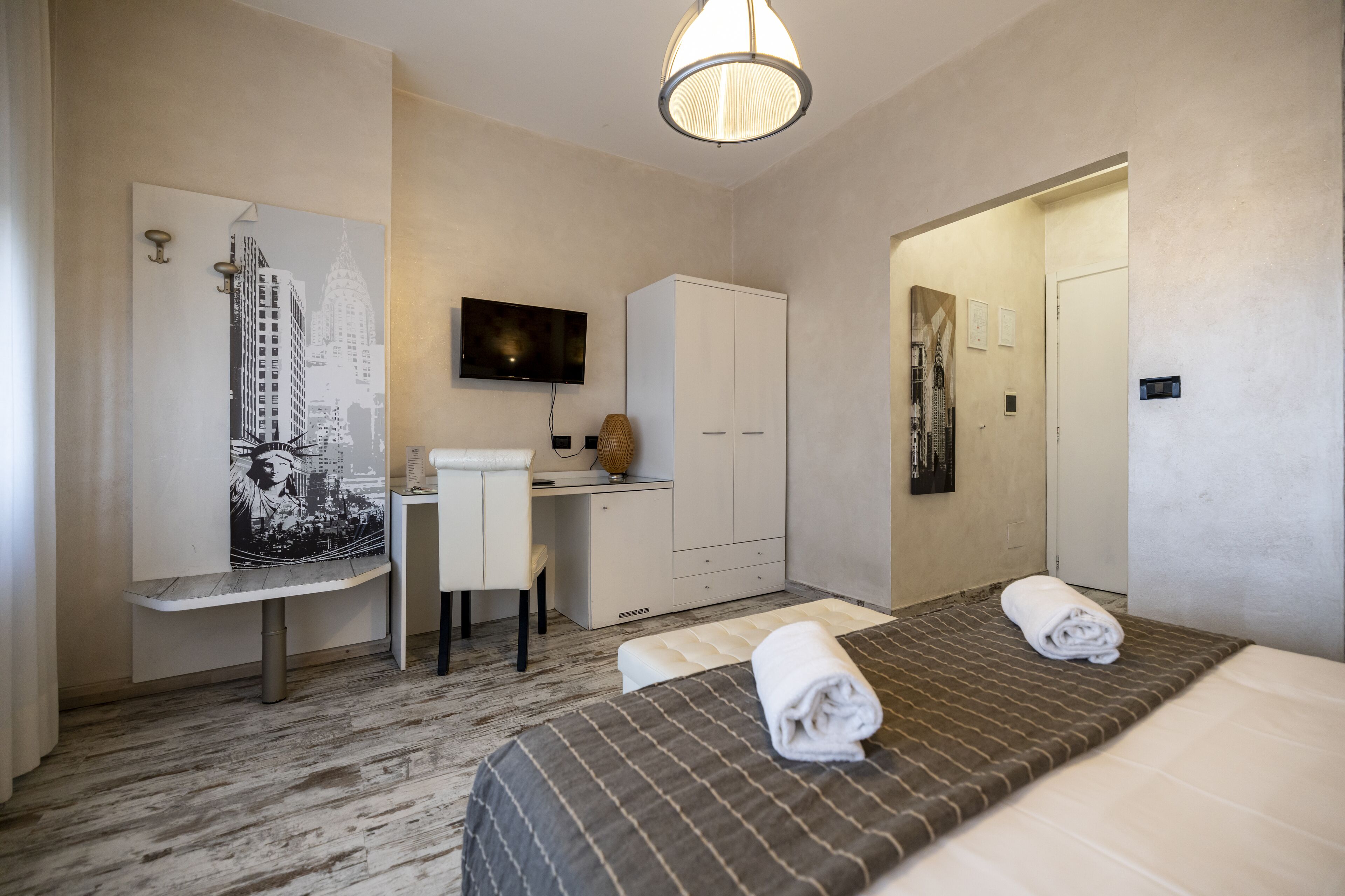 A hotel room with a bed, a desk, a chair, a wardrobe, a television, and a painting on the wall.