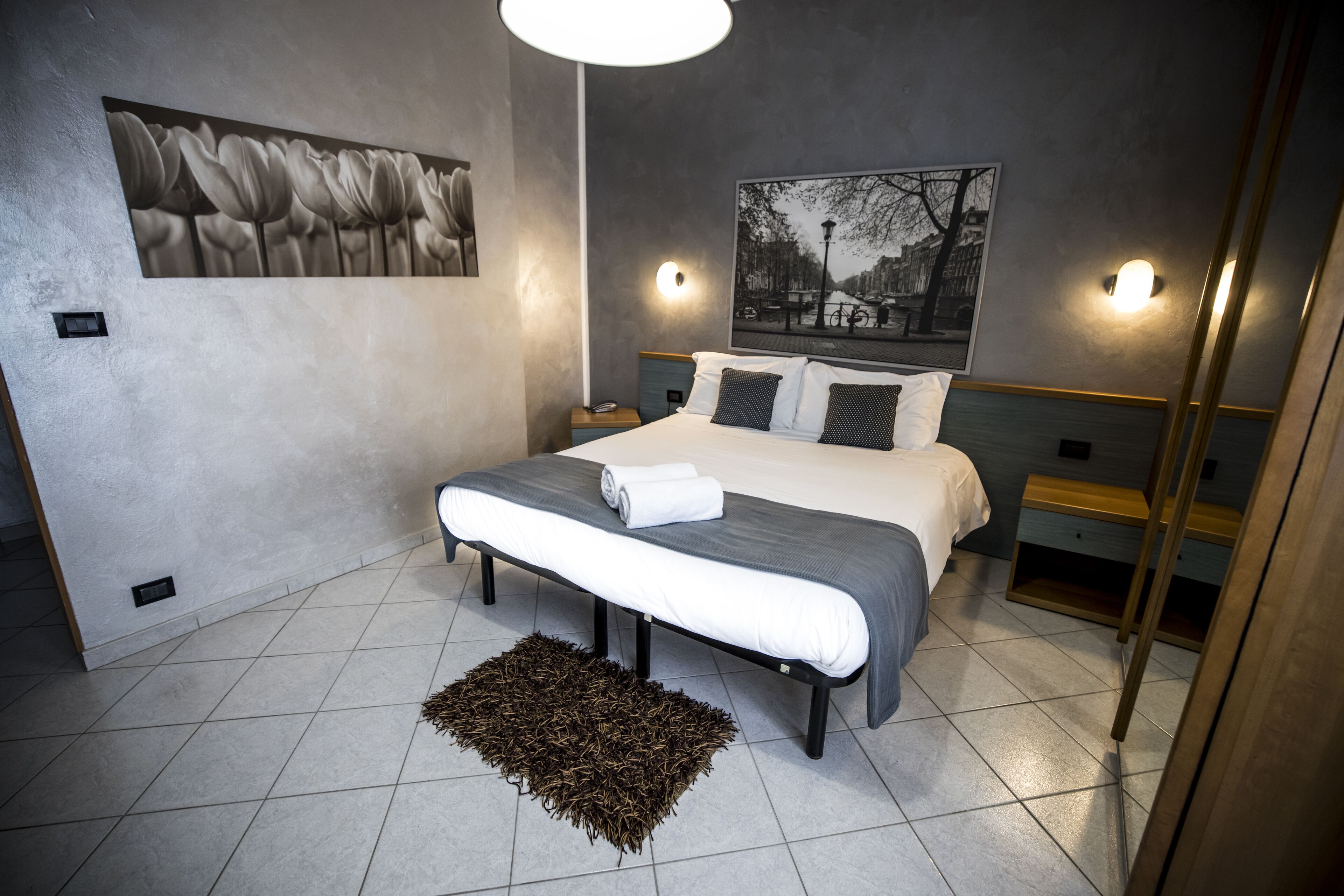 Un dormitorio moderno con una cama grande, dos mesitas de noche, una lámpara de pared y un cuadro con vista a la ciudad.