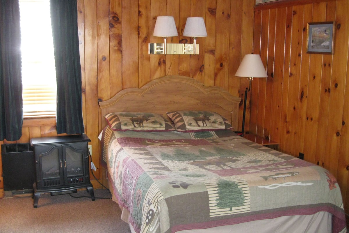 A wooden-paneled room with a bed, two wall-mounted lamps, a heater, and a framed picture on the wall.