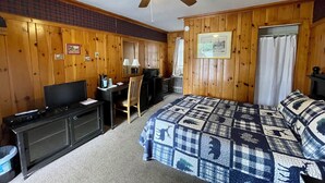 A room with wooden walls, a ceiling fan, a bed with a patterned quilt, a desk with a computer, a TV, and a small kitchenette area. - Mountain Brook Lodge (Wilmington)