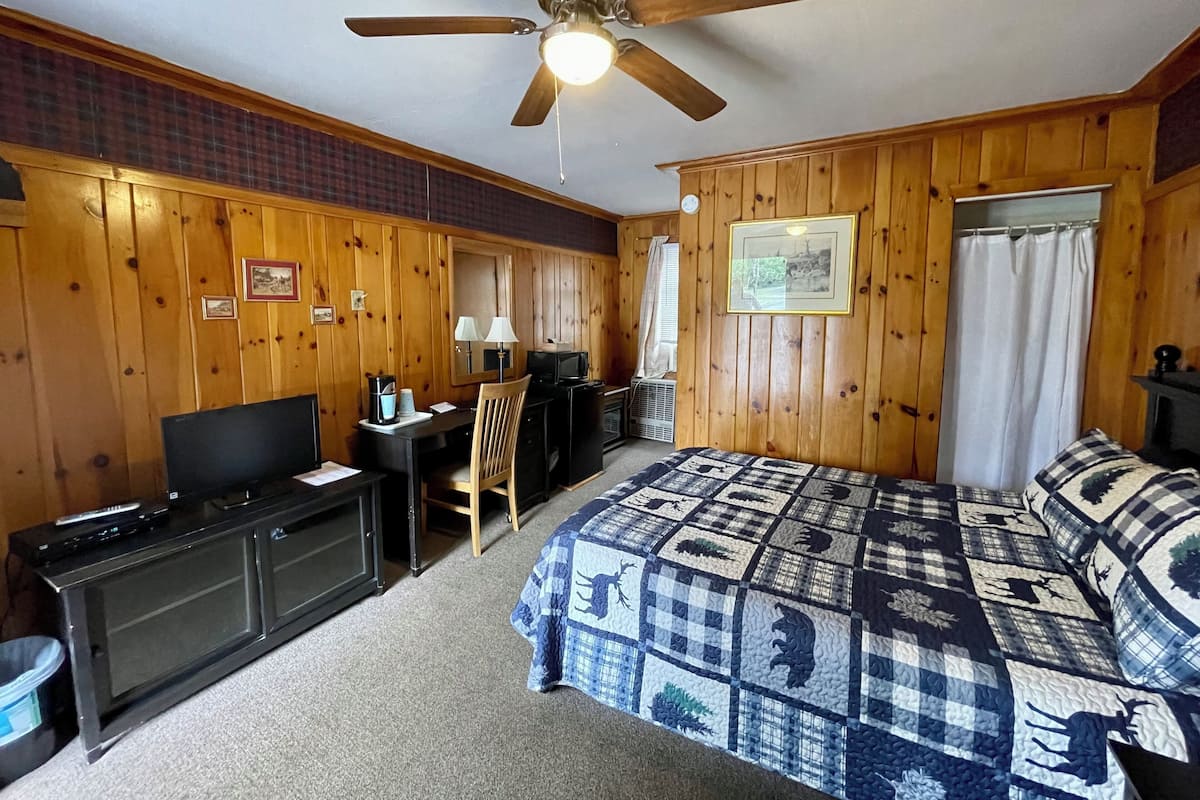 A room with wooden walls, a ceiling fan, a bed with a patterned quilt, a desk with a computer, a TV, and a small kitchenette area.