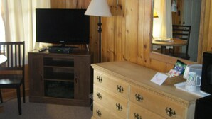 A room with wooden walls, a television on a stand, a dresser with drawers, a lamp, a chair, and a table with a chair. - Mountain Brook Lodge (Wilmington)