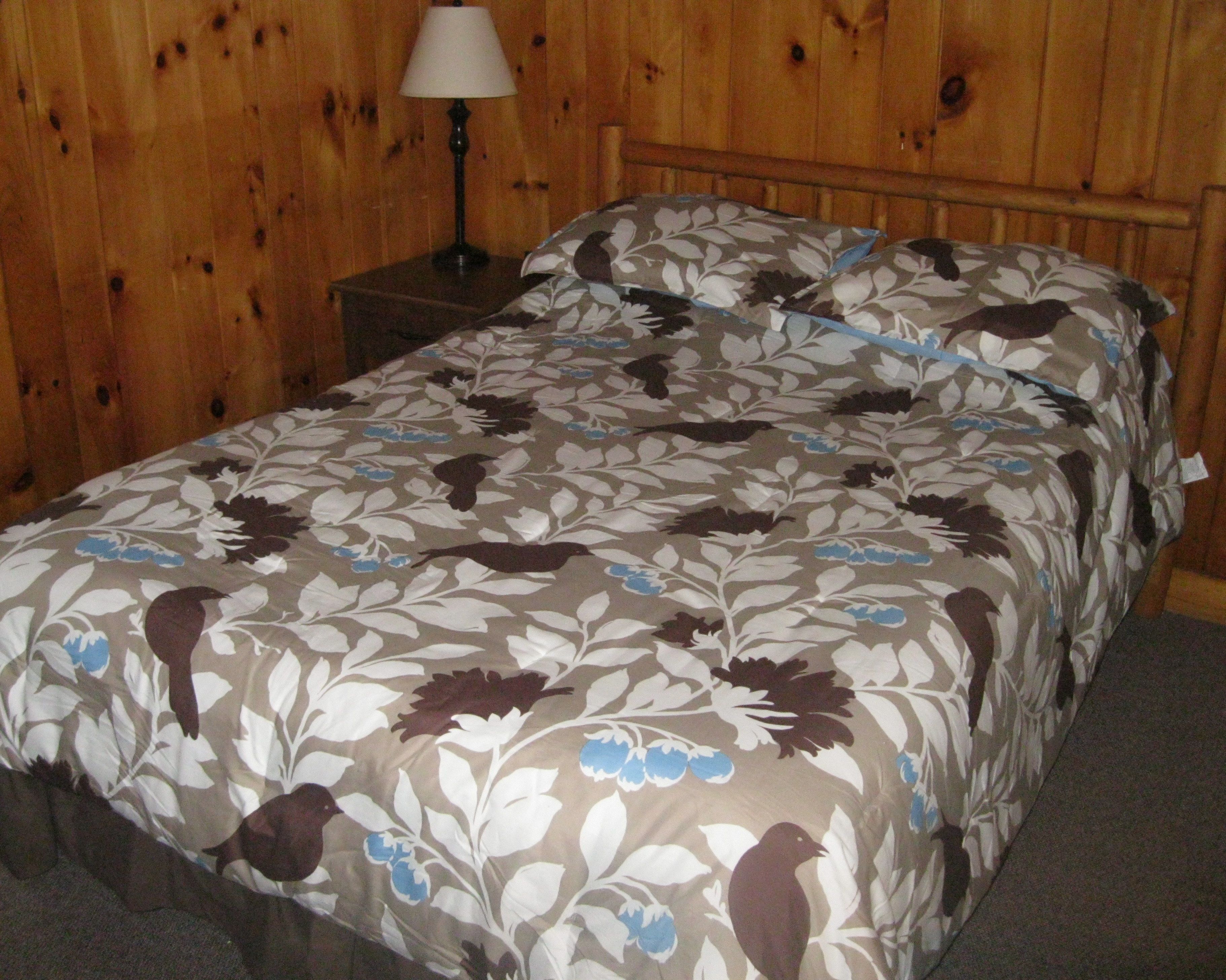 A bed with floral-patterned bedding in a room with wooden walls and a nightstand with a lamp.