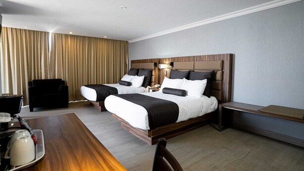 Quarto de hotel com duas camas, uma escrivaninha de madeira, uma cadeira, uma mesinha de cabeceira e um abajur.
