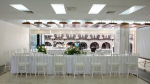 Uma sala de banquetes com mesas e cadeiras brancas, uma varanda com janelas arqueadas e vista para um pátio externo.