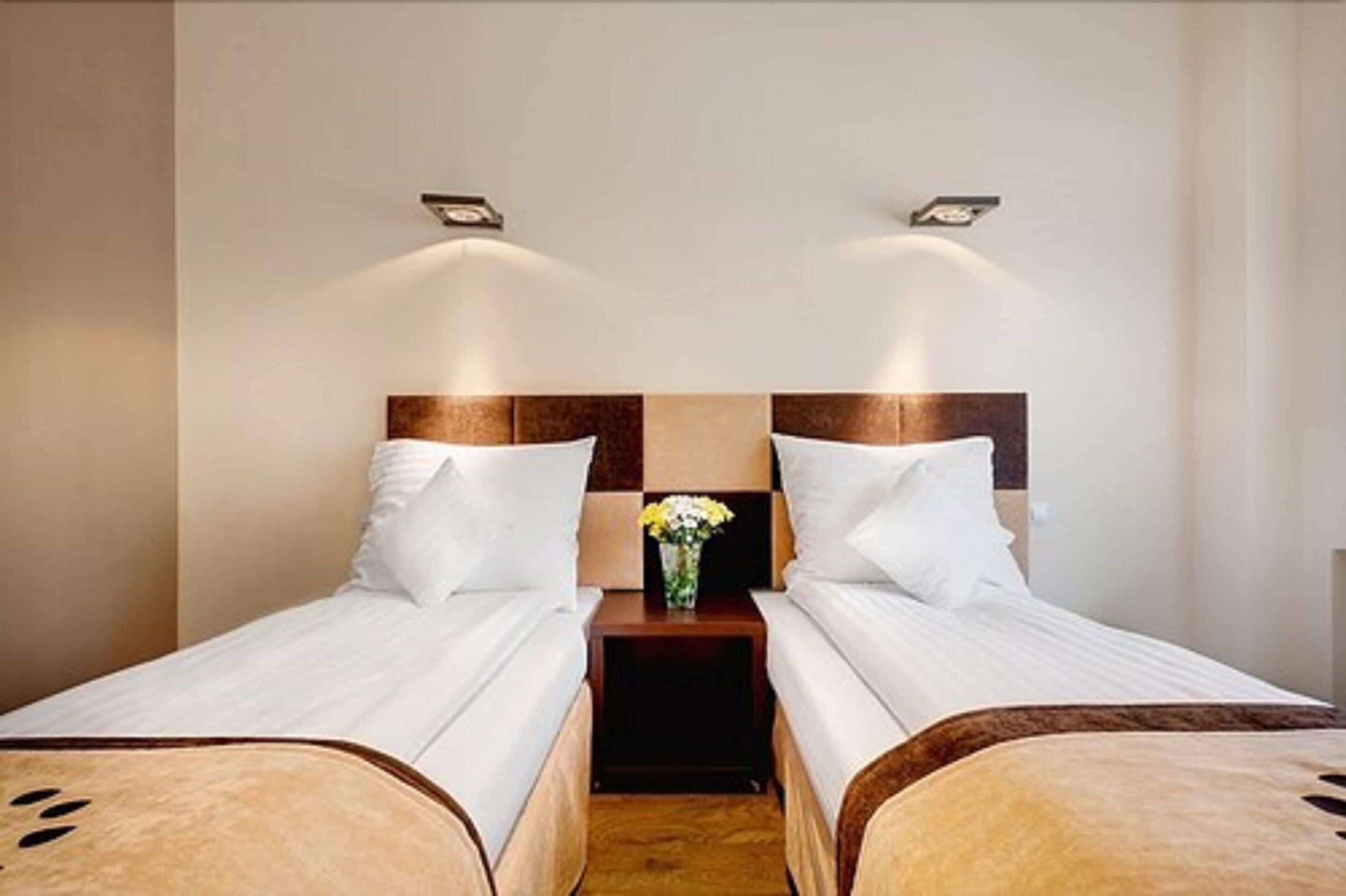 A hotel room with two single beds, a nightstand with a vase of flowers, and wall-mounted lighting.
