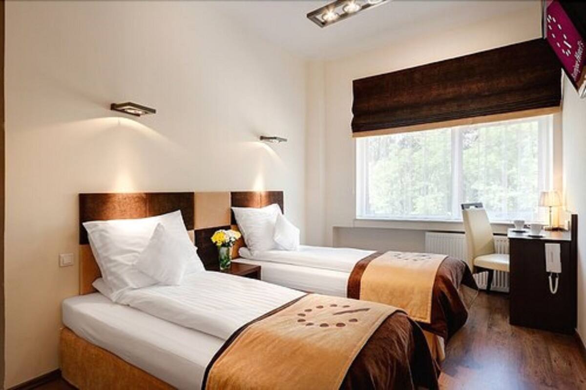 Habitación de hotel con dos camas, un escritorio con lámpara, una silla y ventana con vista a árboles.