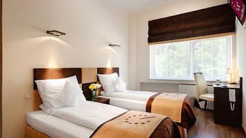 Habitación de hotel con dos camas, un escritorio con lámpara, una silla y ventana con vista a árboles.