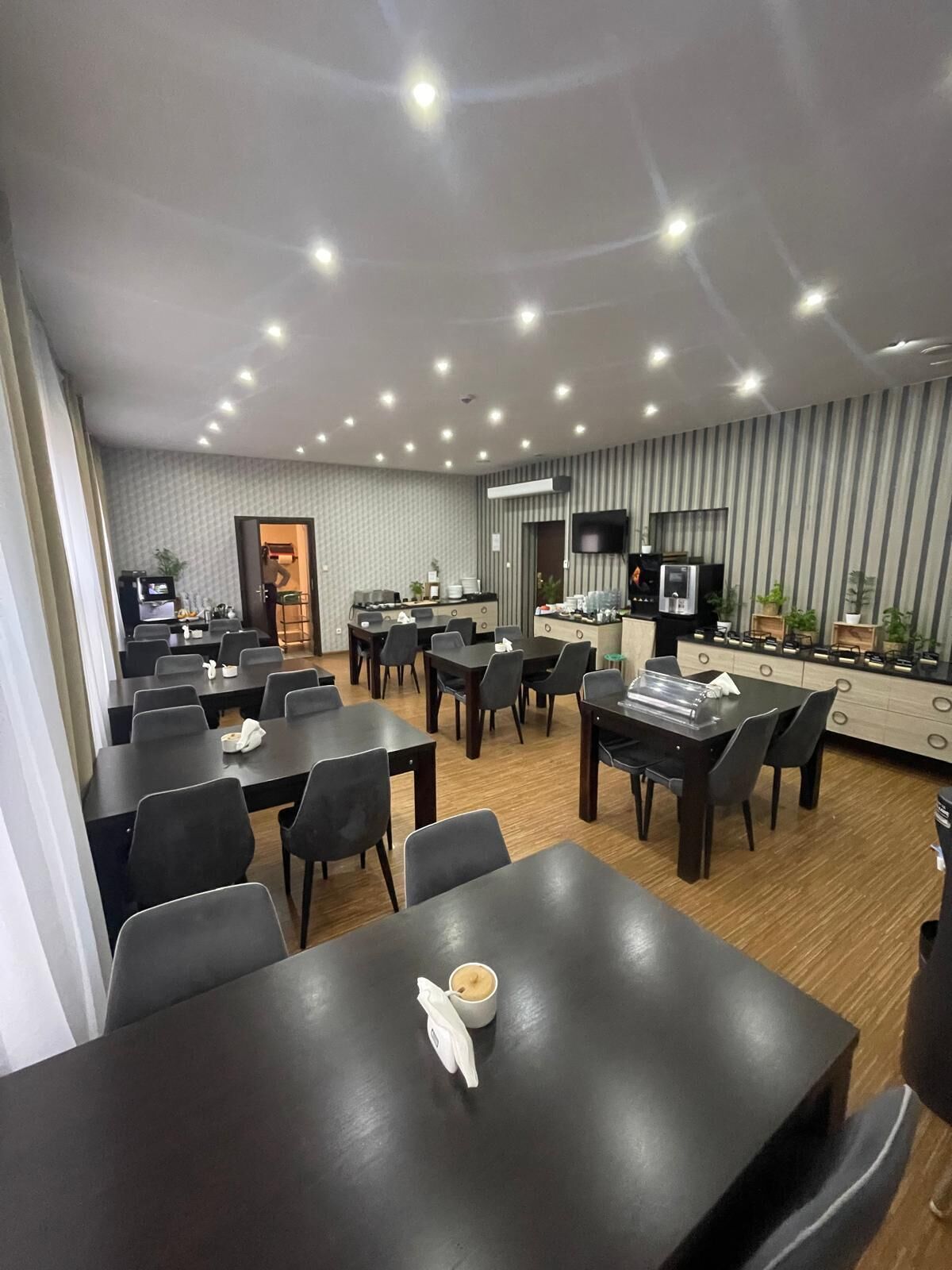A modern dining area with multiple tables and chairs, a kitchen area with appliances, and a television mounted on the wall.