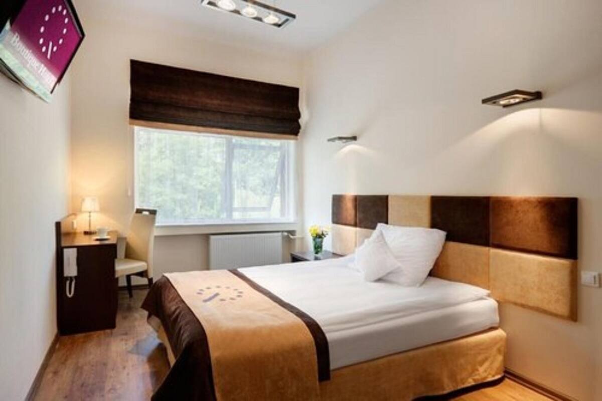 Un dormitorio ordenado con cama, mesita de noche, lámpara y ventana con vistas a zonas verdes.