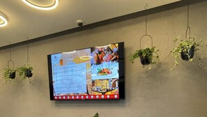 Un espace d’accueil d’hôtel doté d’un téléviseur à écran plat affichant un menu, de deux plantes en pot et d’un plafond orné de luminaires circulaires.