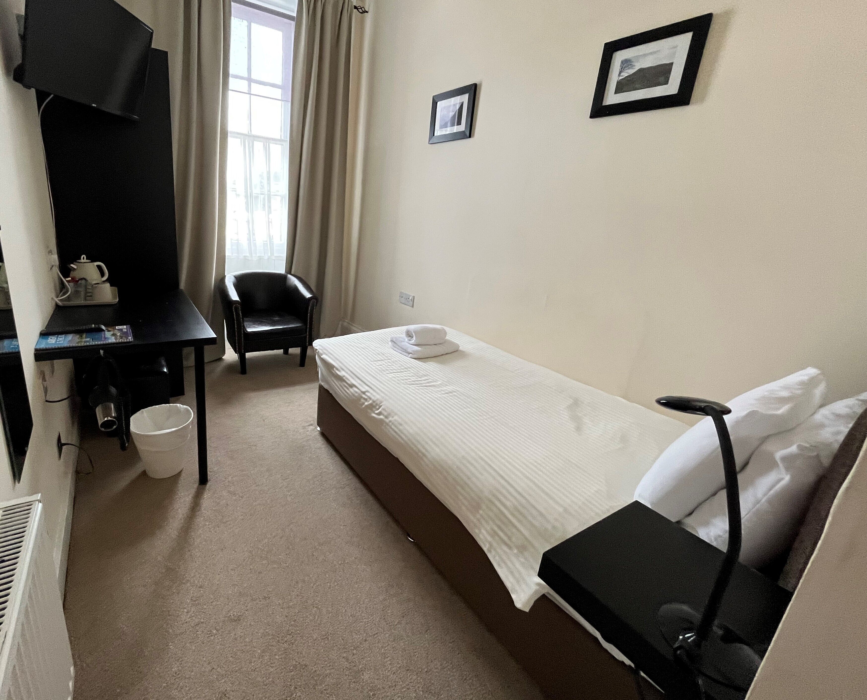 A small, clean hotel room with a single bed, a desk with a chair, a black TV, and a window with curtains.