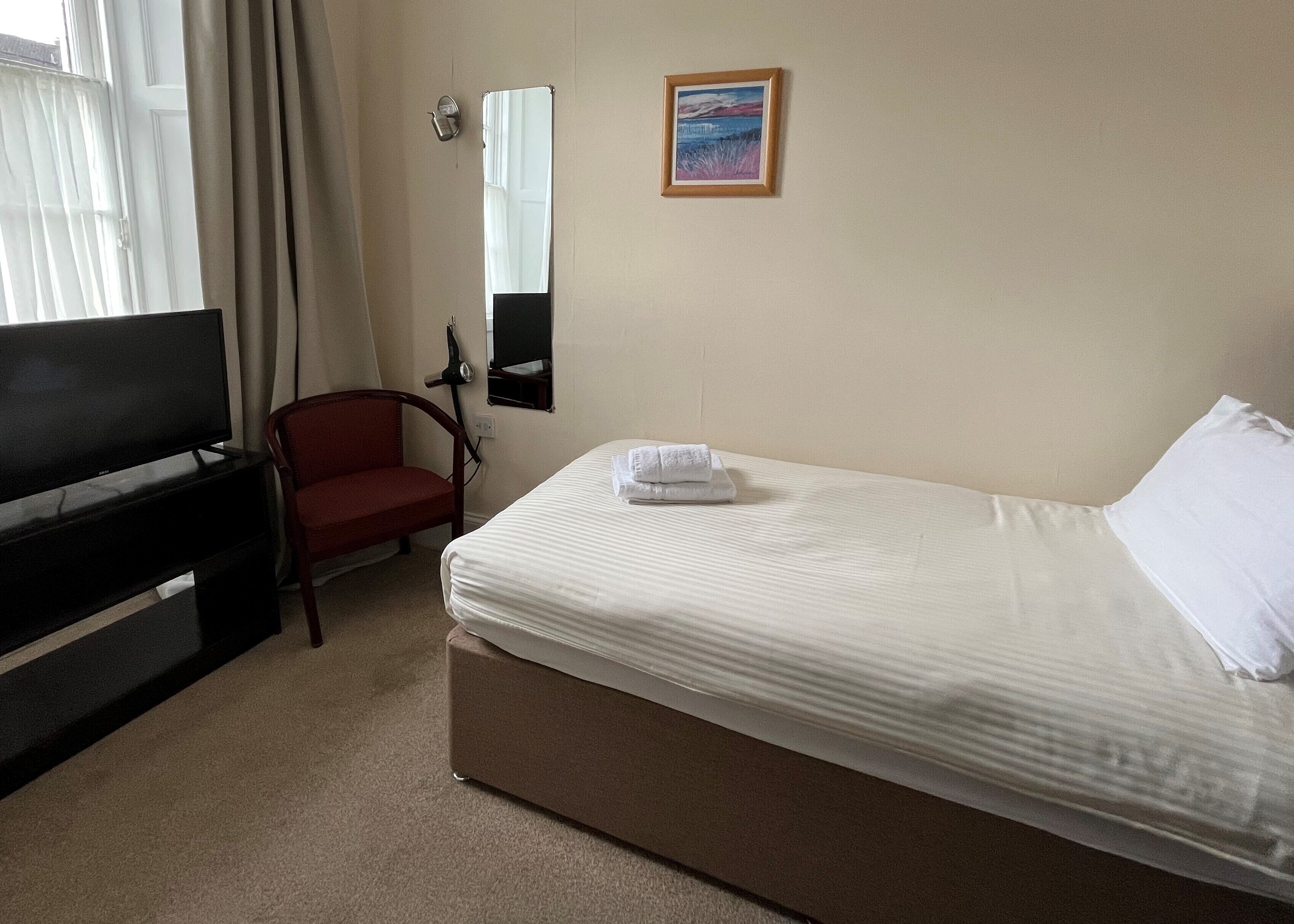 Habitación de hotel con cama, televisión, silla, cuadro y ventana con cortinas.
