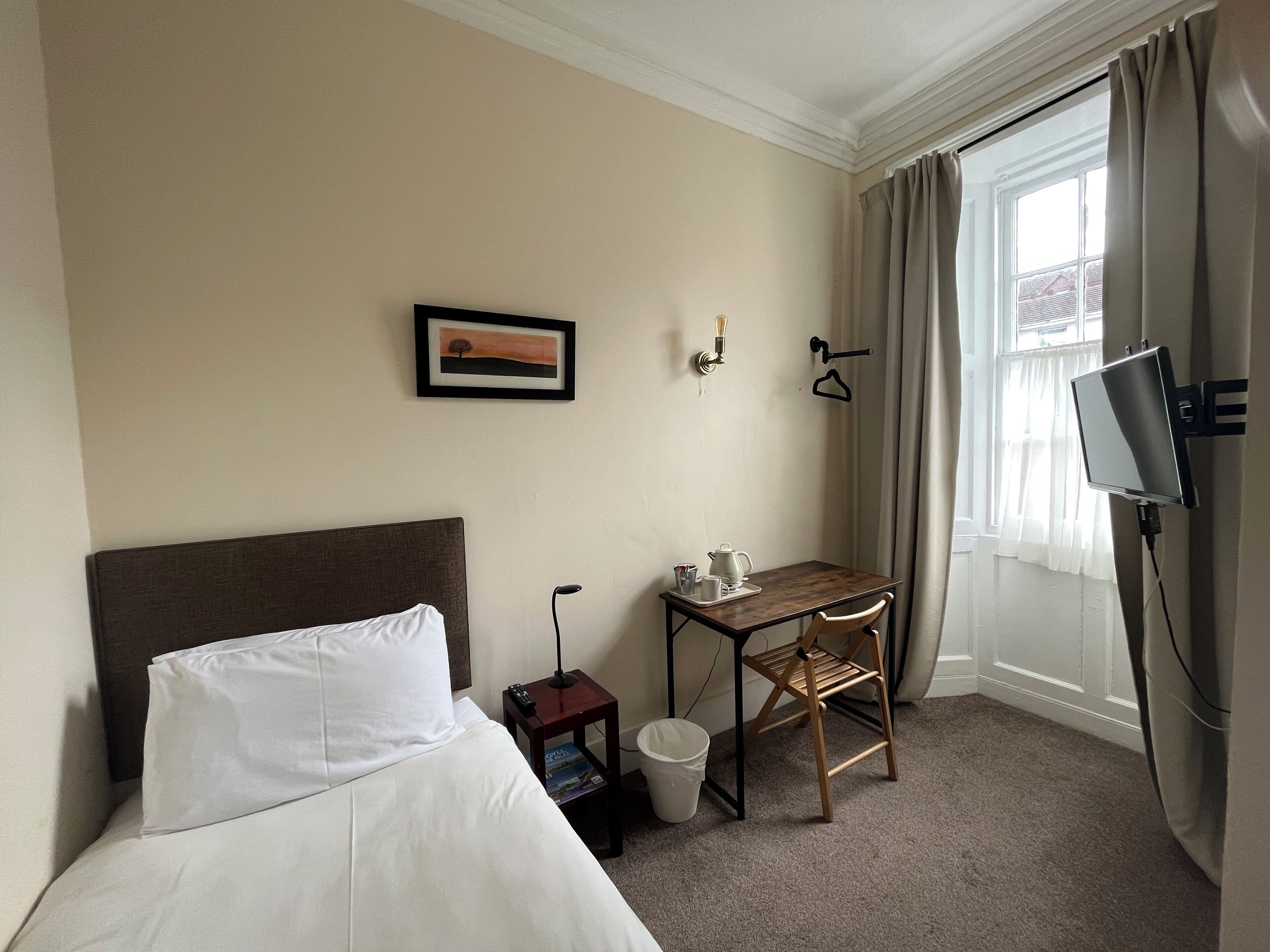 A hotel room with a bed, a small desk with a chair, a television, a picture on the wall, and a window with curtains.