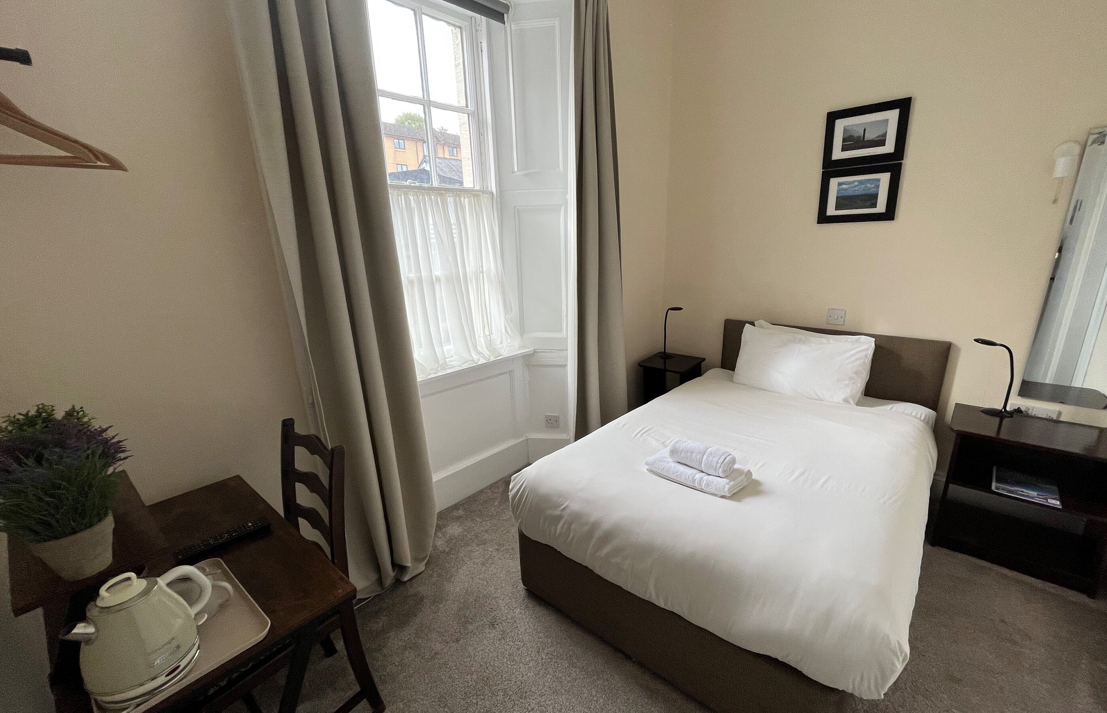 A hotel room with a bed, bedside table, a small table with a kettle and a plant, a window with curtains, and two framed pictures on the wall.