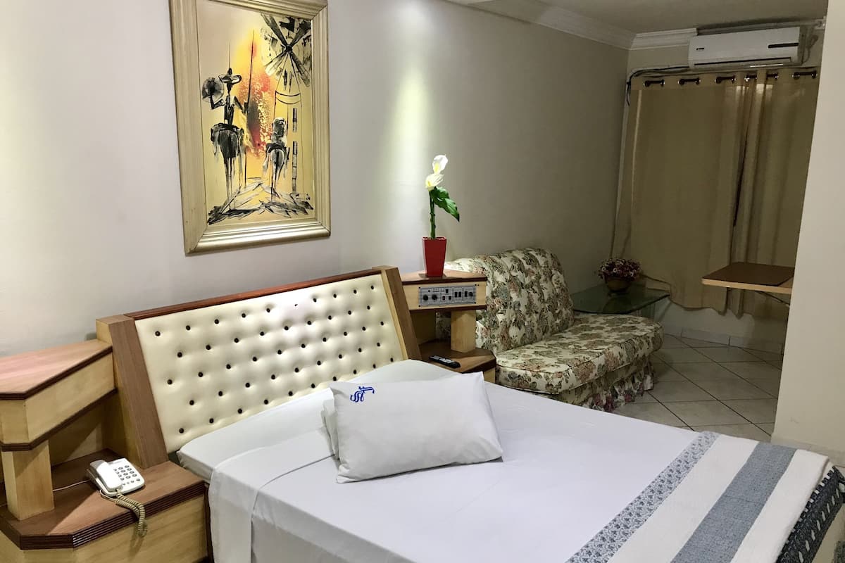 Ein Hotelzimmer mit einem Bett, einem Sessel, einem Nachttisch mit Telefon, einem Bild an der Wand und einer Klimaanlage.