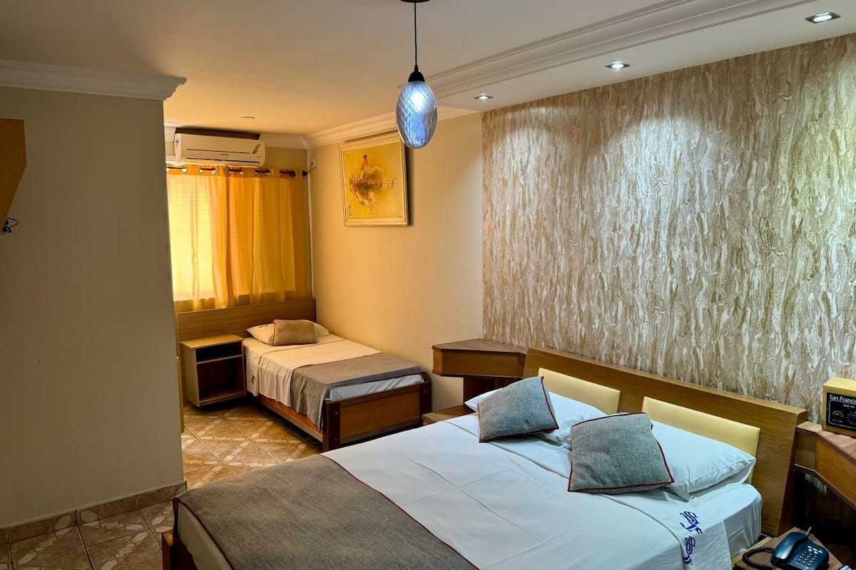 Ein Hotelzimmer mit einem großen Bett, einem kleineren Bett, einem Schreibtisch mit Telefon und einer Wandklimaanlage.