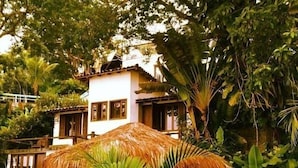 A tropical-style villa with a thatched-roof structure, lush greenery, and a white building in the background. - Pousada Martin Pescador (Búzios)