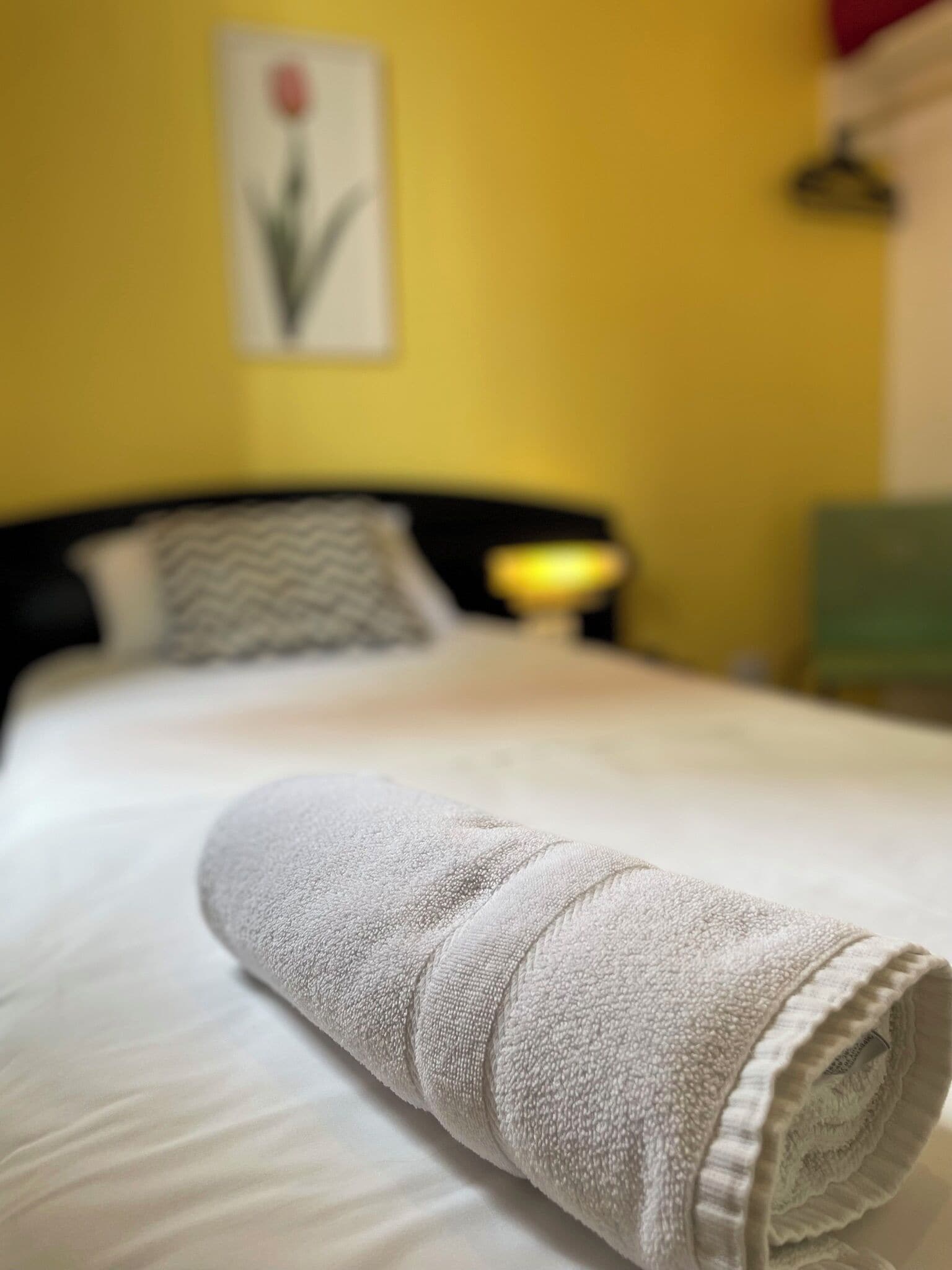 Una cama bien hecha con una toalla blanca enrollada al frente, un cabecero negro y una pared amarilla con un cuadro enmarcado.