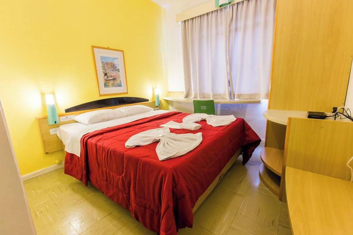 A hotel room with a bed, a window with curtains, a bedside table with a lamp, and a small desk with a chair.