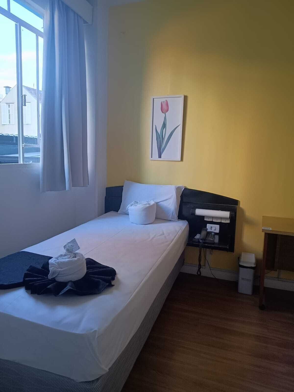 A single bed with white linens, a bedside table with a phone, a small trash bin, and a window with curtains.