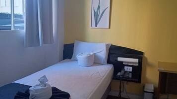 A single bed with white linens, a bedside table with a phone, a small trash bin, and a window with curtains.