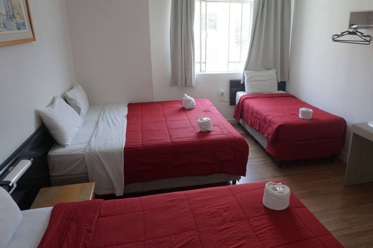 A hotel room with two beds, each with a red bedspread and white pillows, a wooden desk with a fan, and a window with curtains.