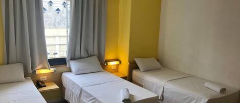 A hotel room with three single beds, a window with curtains, a bedside table with a lamp, and a wall-mounted light.