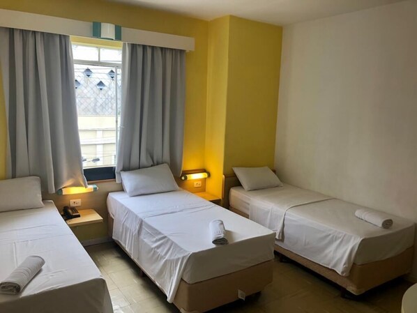 Habitación de hotel con tres camas individuales, una ventana con cortinas, una mesita de noche con lámpara y una lámpara en la pared.