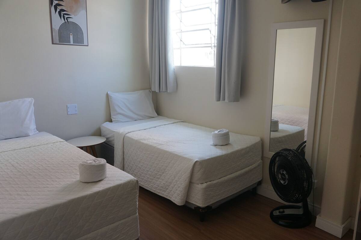 A hotel room with two beds, a small round table, a desk, a chair, a standing fan, and a window with curtains.