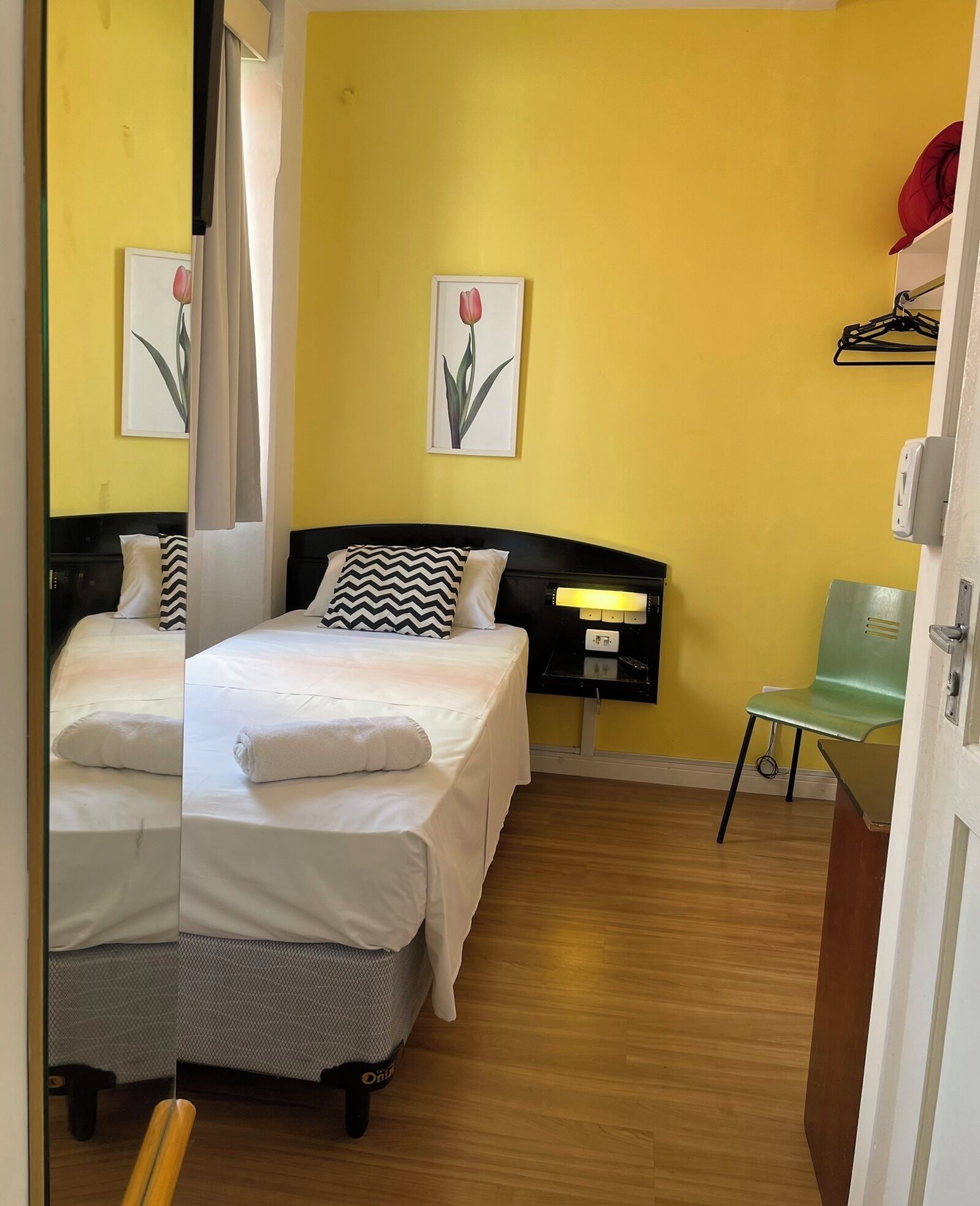 A hotel room with two beds, a bedside table with a lamp, a chair, and artwork on the wall.