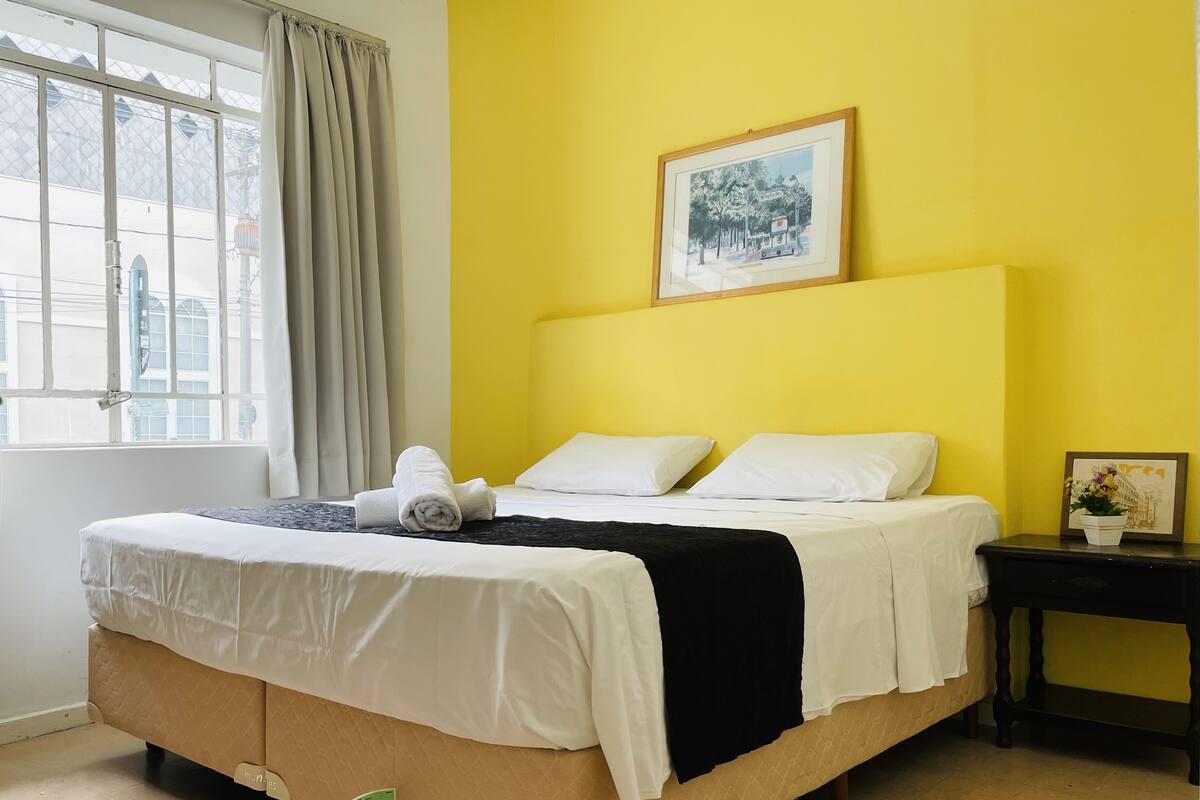 A bed with white linens, a black blanket, and a rolled towel. There is a framed picture on the wall above the bed.