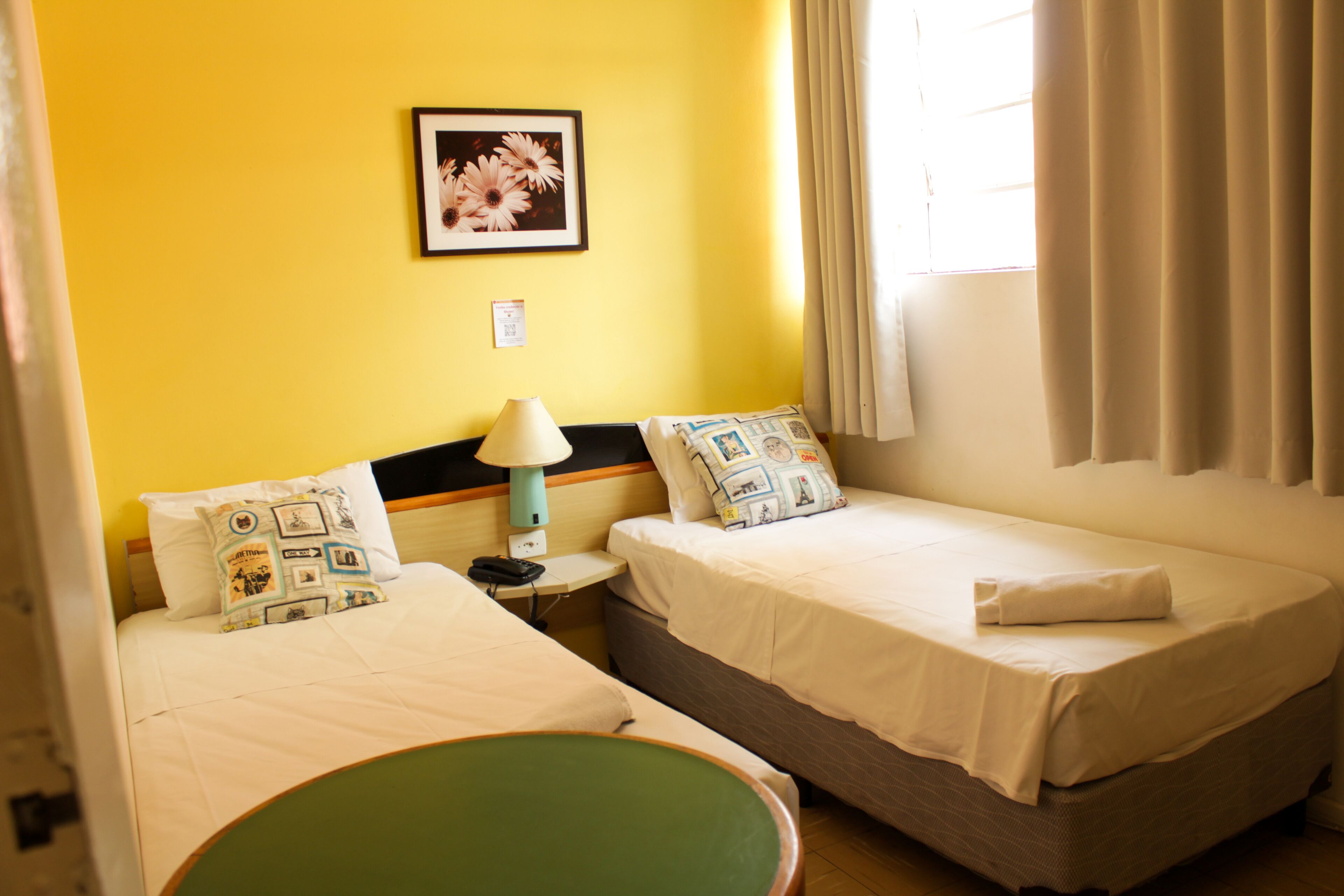 Habitación de hotel con dos camas, una mesita de noche con lámpara, un cuadro enmarcado en la pared y una ventana con cortinas.